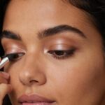 Mulher adulta aplicando sombra clara na pálpebra e iluminador no canto interno dos olhos