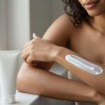 Mulher de pele morena e cabelo escuro espalha protetor solar no antebraço em um banheiro iluminado por uma janela