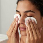 Mulher de olhos fechados apoia disco de algodão umedecido nos cílios, com cotonetes e frasco sem rótulo ao lado